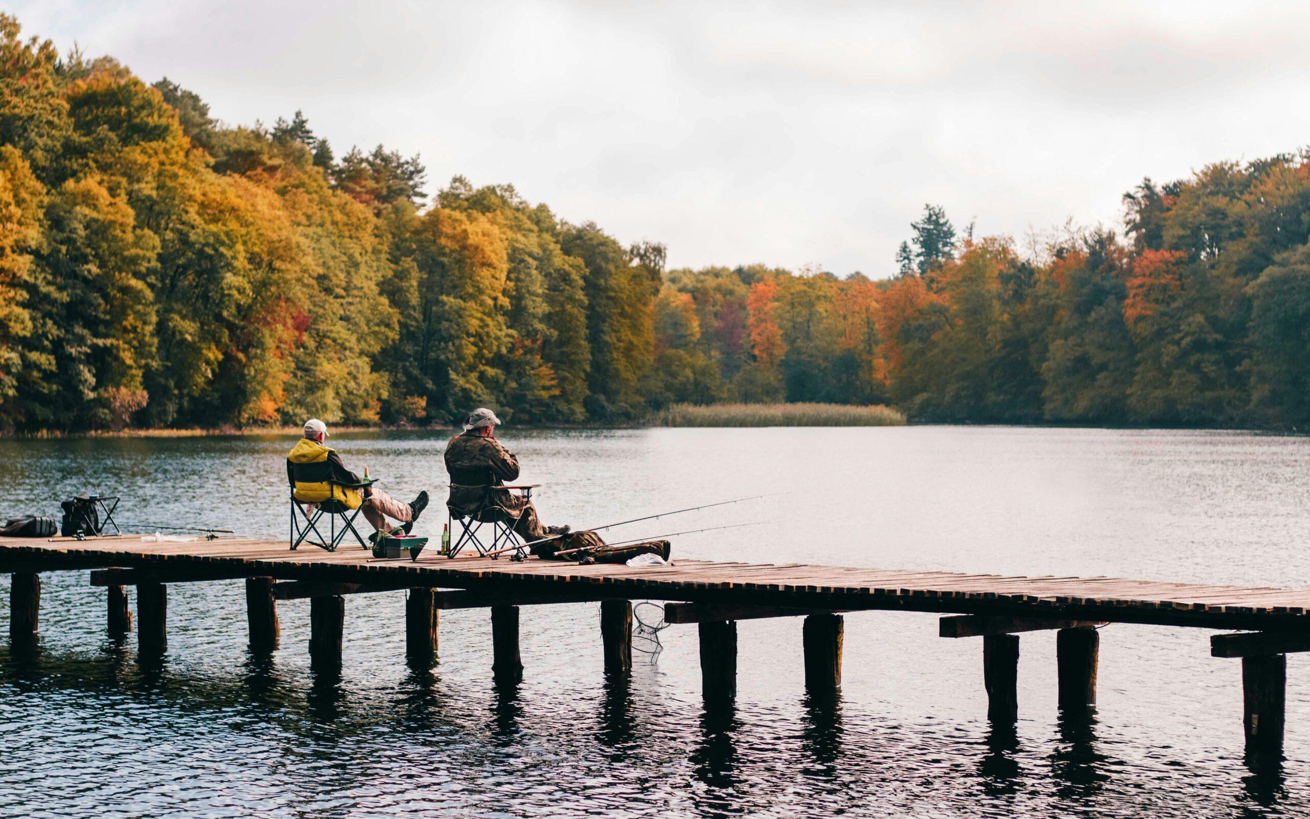 Two people fishing from a wooden dock on a lake framed by autumn trees
