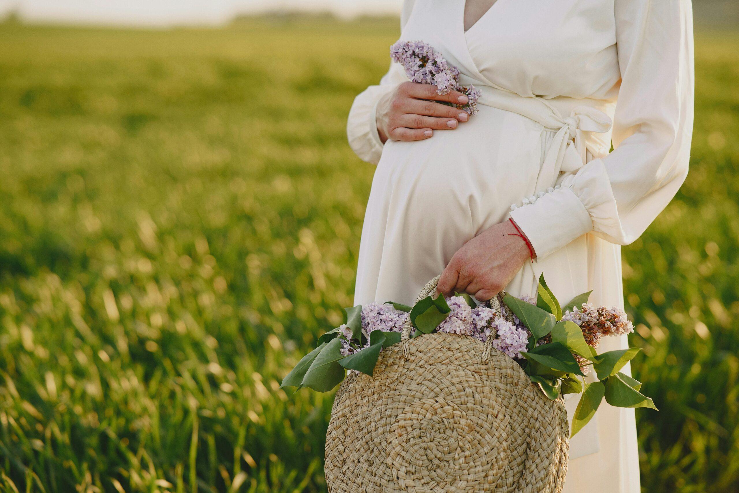 Expecting mother in a white dress holding a basket of wildflowers in a meadow on her babymoon