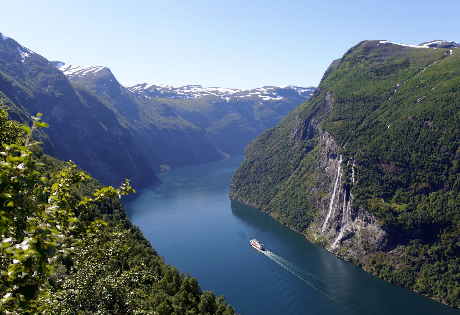 Cruise ship sailing through Norway's Geirangerfjord between steep green mountains and a cascading waterfall