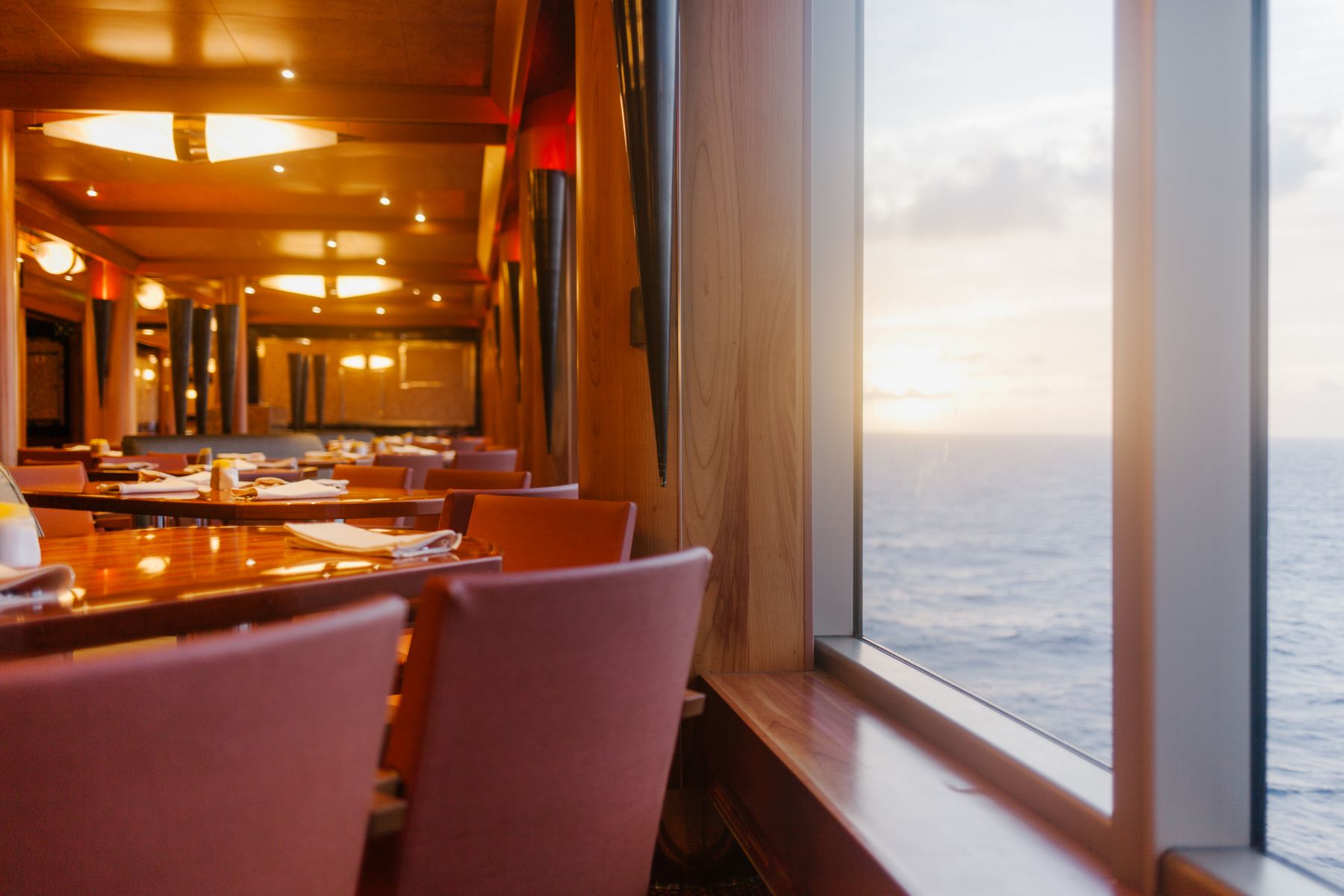 Wood paneled dining room aboard a premium cruise ship with floor to ceiling windows and ocean views at sunset